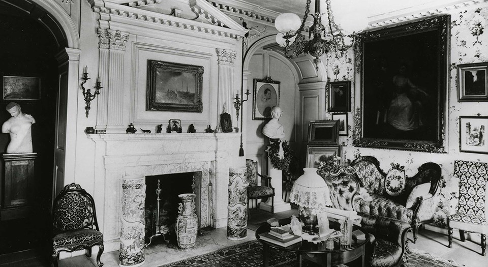 Black and white photograph of room with fireplace, couches and chairs, a gas chandelier, and many paintings on the walls