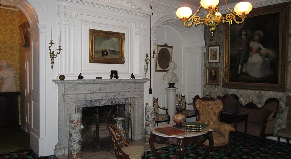 Black and white photograph of room with fireplace, couches and chairs, a gas chandelier, and many paintings on the walls
