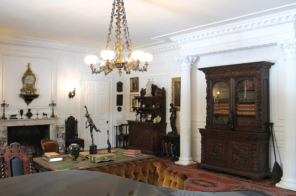 Black and white photograph of Victorian interior with ornate furniture including bookcase and table piled with books.