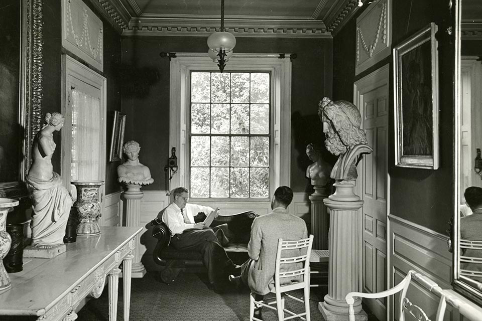 Two men seated in front of a large window, surrounded by statuary.