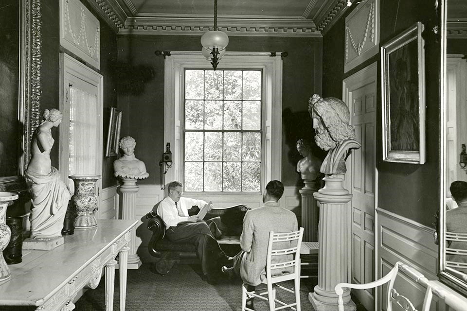 Two men seated in front of a large window, surrounded by statuary.