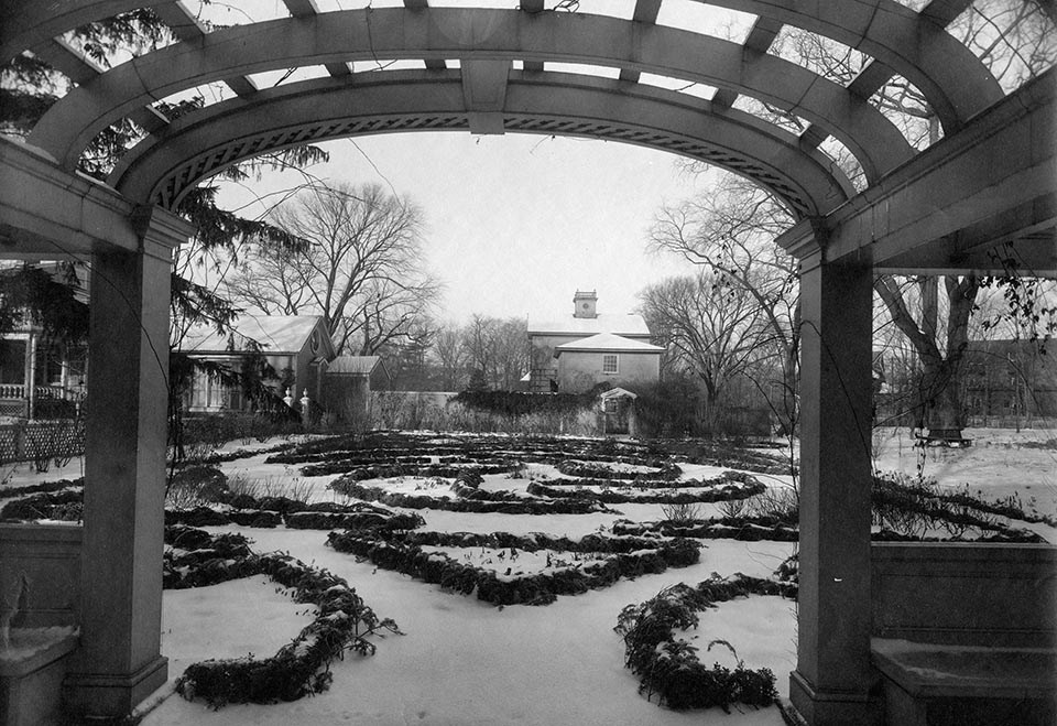 Black and white photograph of garden beds covered in snow framed by arch of pergola structure