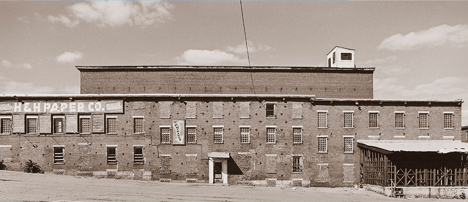 A brick building with the signage "H & H Paper Co." The windows are in various states from being boarded up to bricked in. The rooftop is flat.