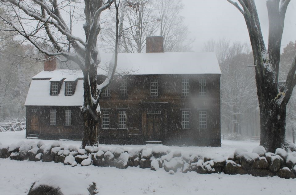 A wooden house with two and a half stories covered in snow. Two large trees tower in front of the building and a stone fence marks the boundary of the road.