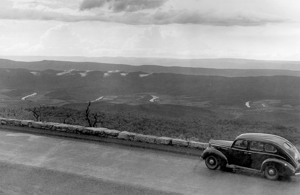 A black and white photograph of an overlook with a vehicle and valley in the distance.