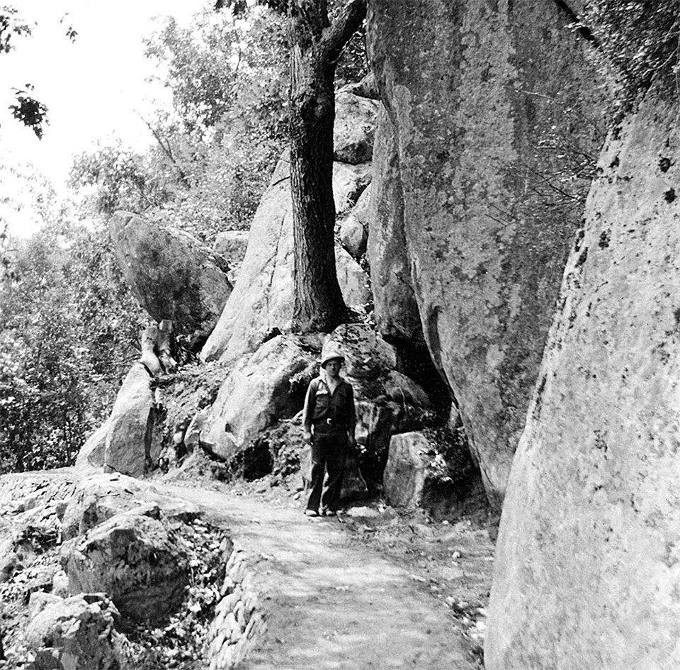 A black and white photograph of a person standing on a trail along a mountainside.