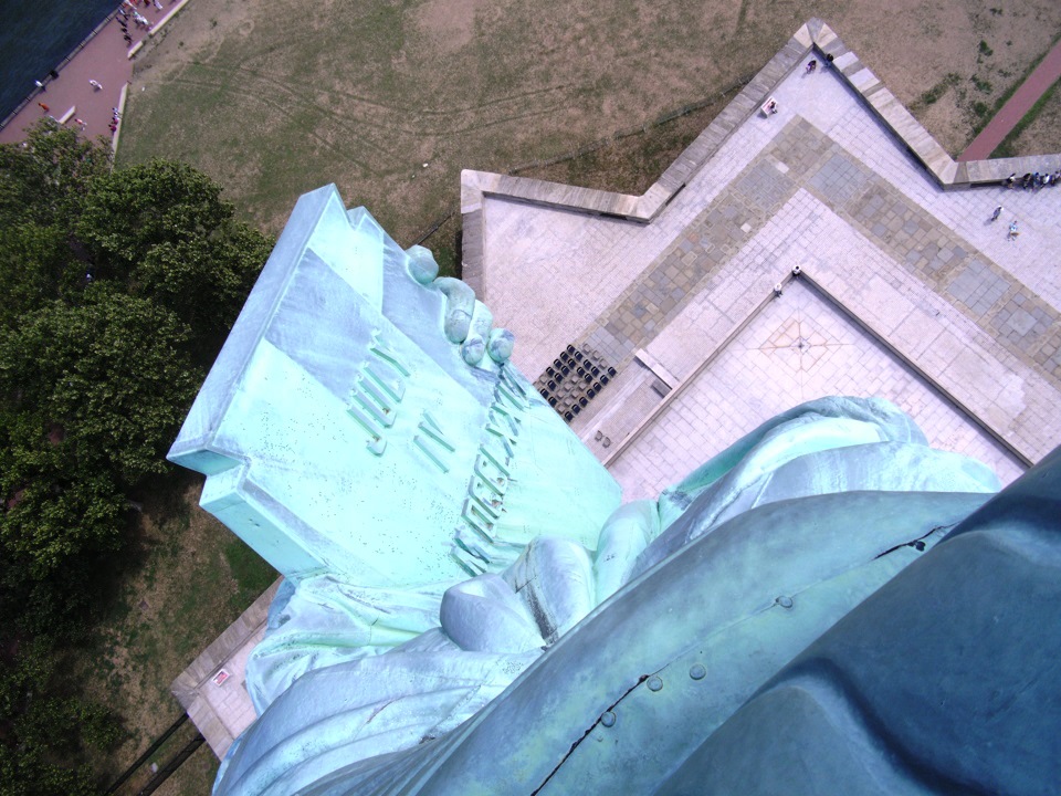 The Statue of Liberty's tablet looking out and down the windows of the crown. It's a sunny day and the grass and trees are green.