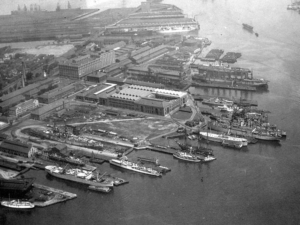 Photograph of roadways, piers, buildings, dry docks, and ships at a naval shipyard.
