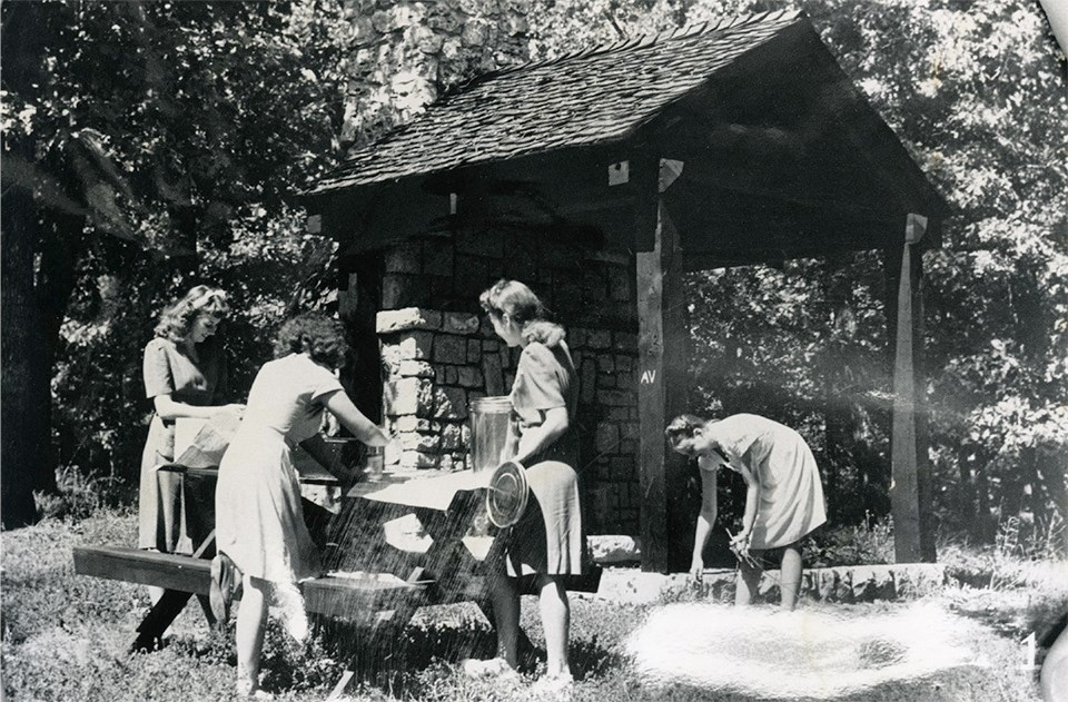 Historic image of four young women in dresses setting up a picnic at a table near a covered shelter.