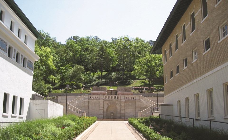 Illustration of a wide stone path with a red brick building on the right. Path leads to an elaborate stone stairway with lamp posts leading up to a hillside with trees and a stone building with columns.