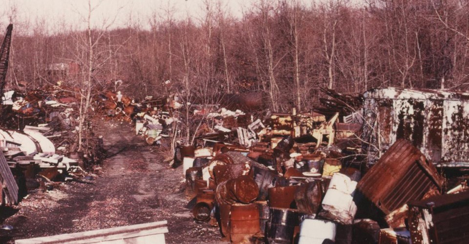 An unpaved road through tall stacks of metal and wooden debris mixed with old tires and rusting drums. Bare trees line the edges of the dump.