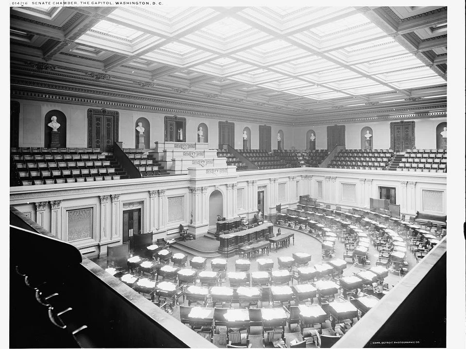 Senators seated in the Senate chamber. Dark wood desks form a semi-circle around the room
