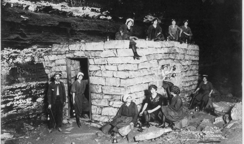 A group of people sit and stand around a rectangular stone hut inside a cave in 1912.