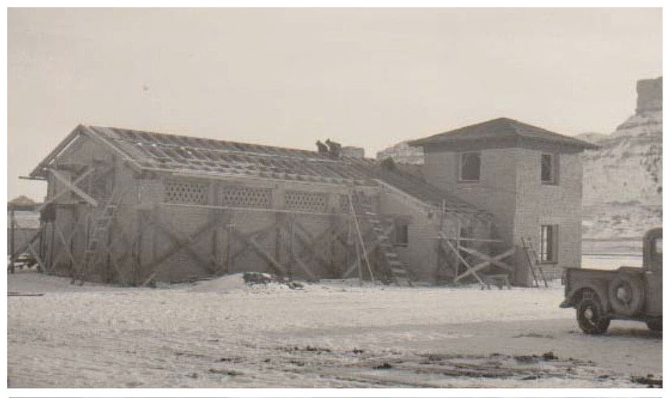 Historic image of a long adobe structure with a tower at one end under construction.