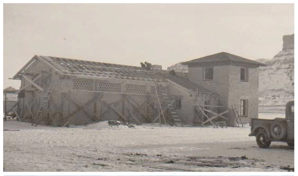 Historic image of a long adobe structure with a tower at one end under construction.