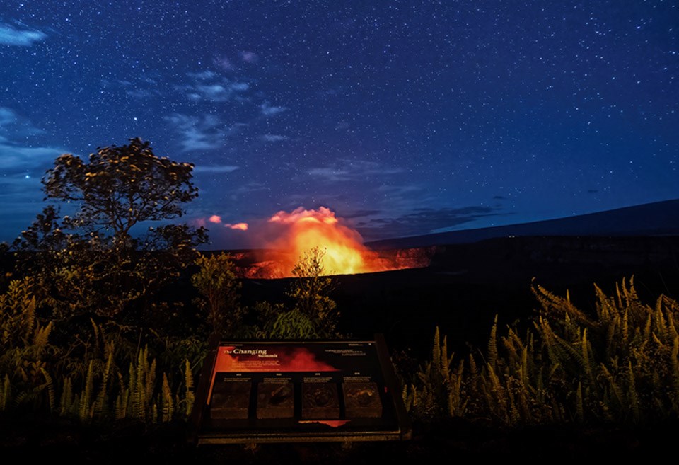 A glowing volcanic crater with a wayside exhibit in the foreground underneath a starry sky