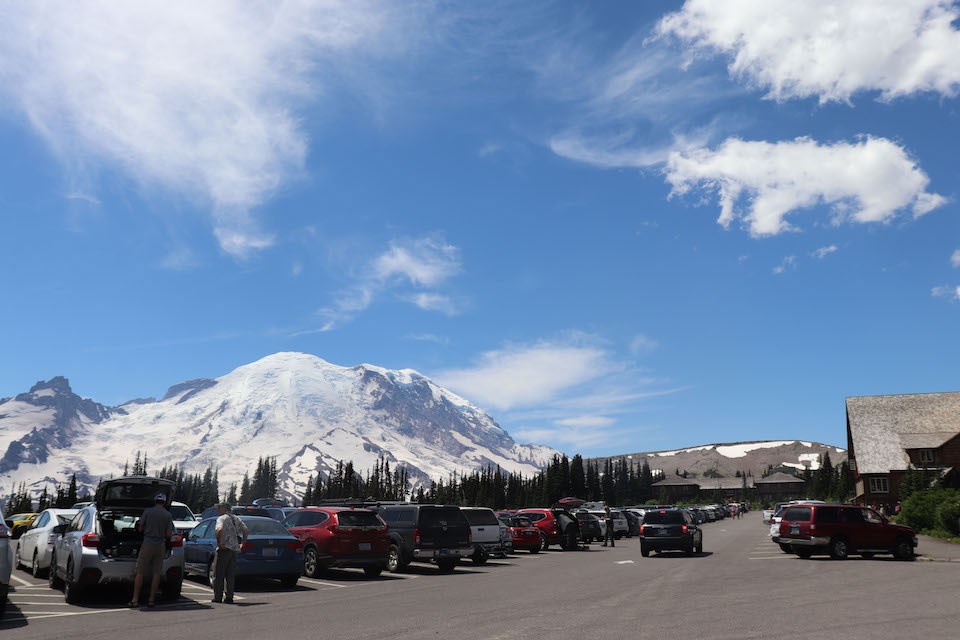 A parking lot full of cars and people with a sunny sky and Mount Rainier in the back.