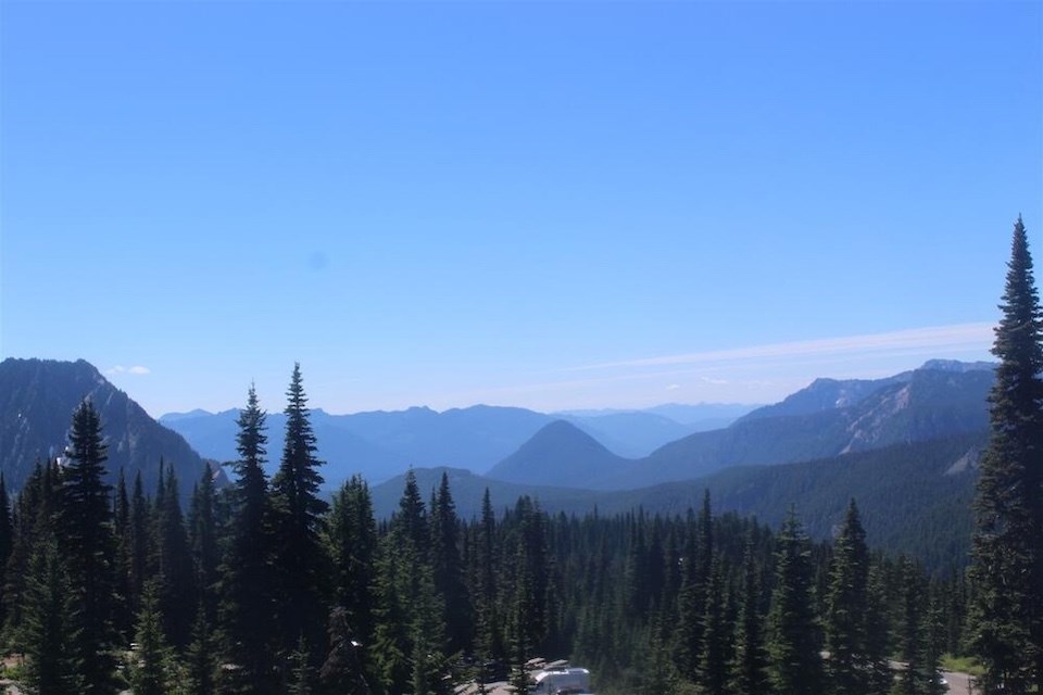 Webcam still showing a series of forested mountain peaks with a clear blue sky.
