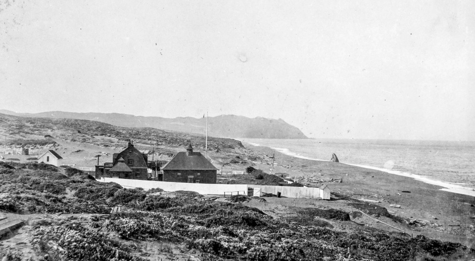A black and white photo of several wooden structures adjacent to an ocean beach on the right with rugged headlands rising in the distance.