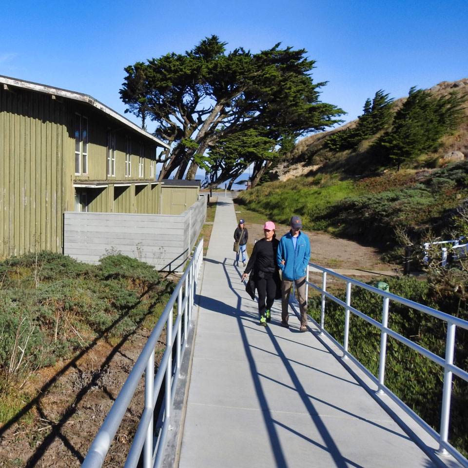 A black and white photo of a snowy pathway with tricycle tracks and footprints leading to a two-story house on the left. A man stands near the near corner of the house.