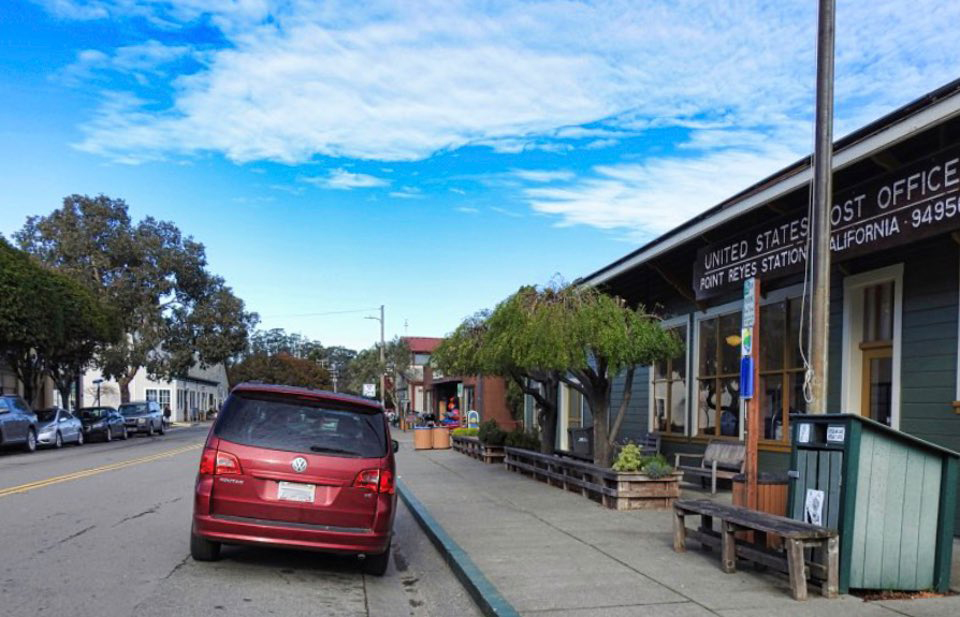 A black and white photo of people milling about a steam locomotive that is parked on tracks adjacent to a one-story-tall train station on the right and a dirt road on the left.