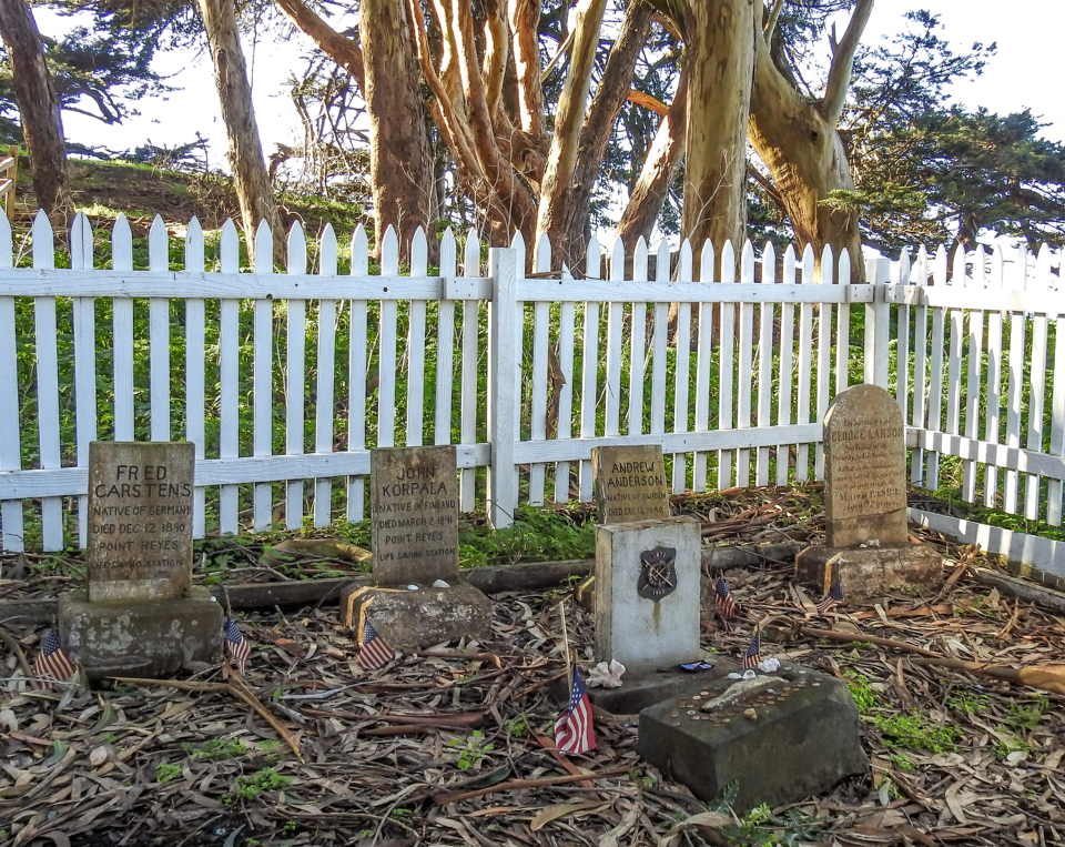 A black and white photo of four grave markers surrounded by a picket fence and trees.