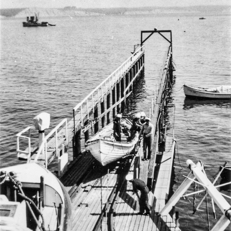 A black and white photo of uniformed men lowering an open-topped surfboat down a rail into a bay.