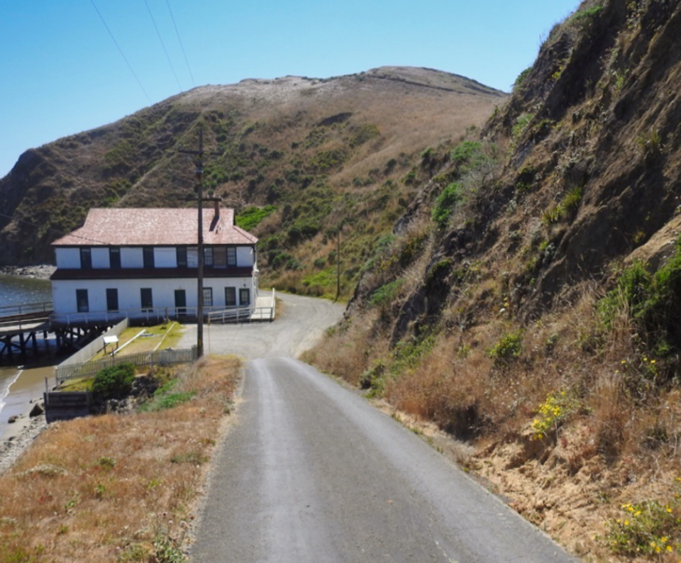 A black and white photo of men clearing landslide debris from a road that leads down to some bay-side buildings. Steep bluffs rise on the right and in the background.