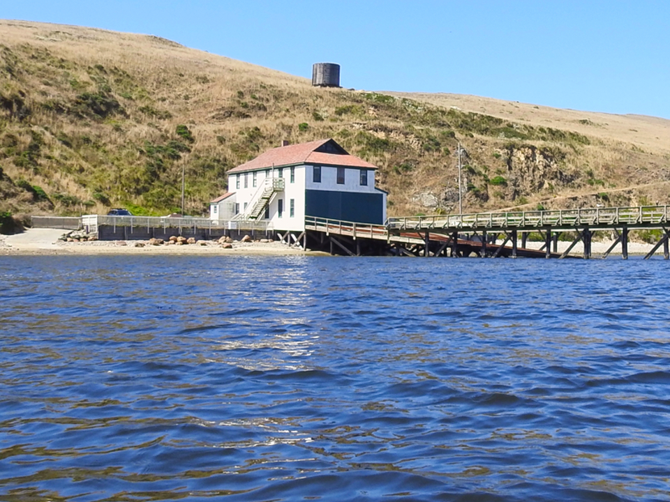 A black and white photo of a man in a plaid, long-sleeved shirt rowing a boat away from a white-sided, two-story boathouse.