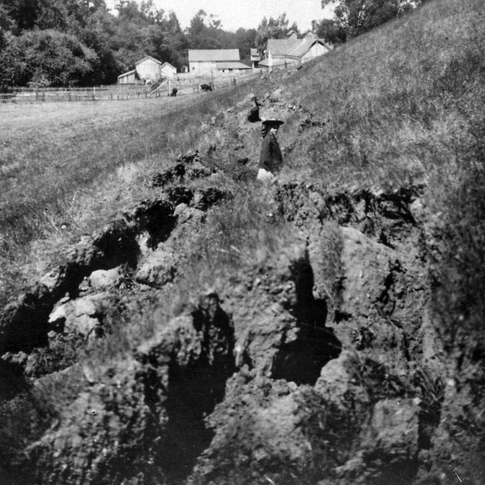 A black and white photo of a woman standing in a surface rupture, where the soil has been turned over, with a barn in the background.