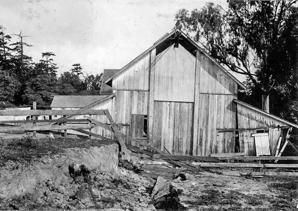 A black and white photo of a barn that is tilted slightly to the right and a broken fence in the foreground.