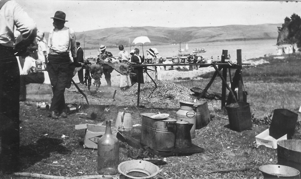 A black and white photo of a cookout attended by men and women on a beach as boats float in the bay in the background.