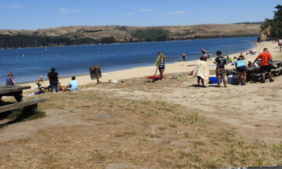 A black and white photo of a cookout attended by men and women on a beach as boats float in the bay in the background.