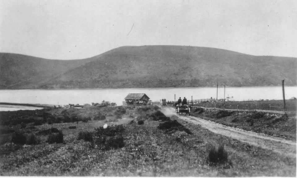 A black and white photo of horse-drawn wagons traveling along a dirt road that leads to a building and a dock on a body of water with hills in the background.
