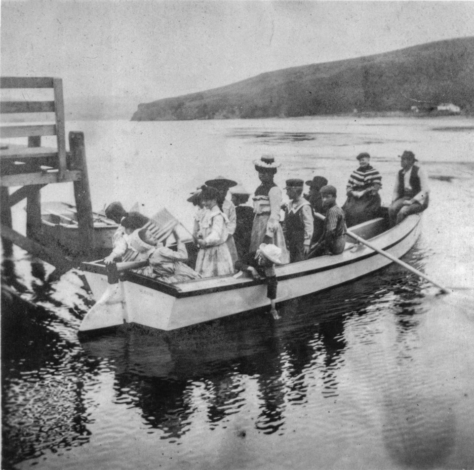 A black and white photo of thirteen people in a rowboat adjacent to a dock on the left. Low headlands rise above the far end of the bay.