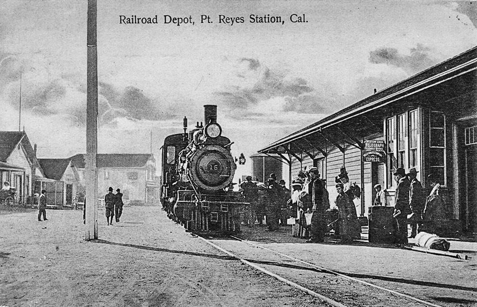 A black and white photo of people milling about a steam locomotive that is parked on tracks adjacent to a one-story-tall train station on the right and a dirt road on the left.