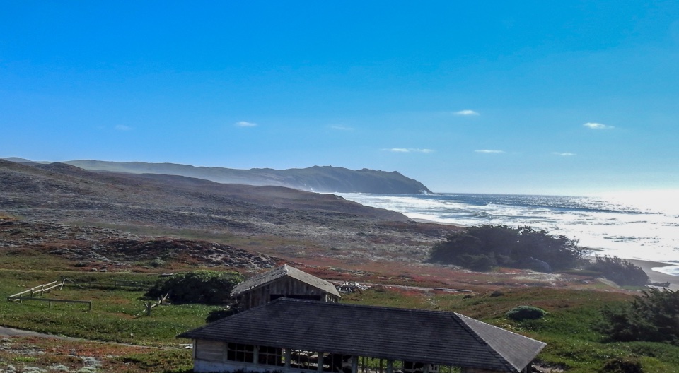A black and white photo of several wooden structures adjacent to an ocean beach on the right with rugged headlands rising in the distance.