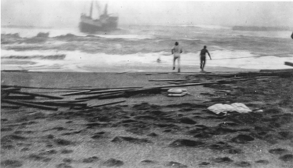Black and white photo of two men on a beach with a ship caught in heavy surf very close to shore.