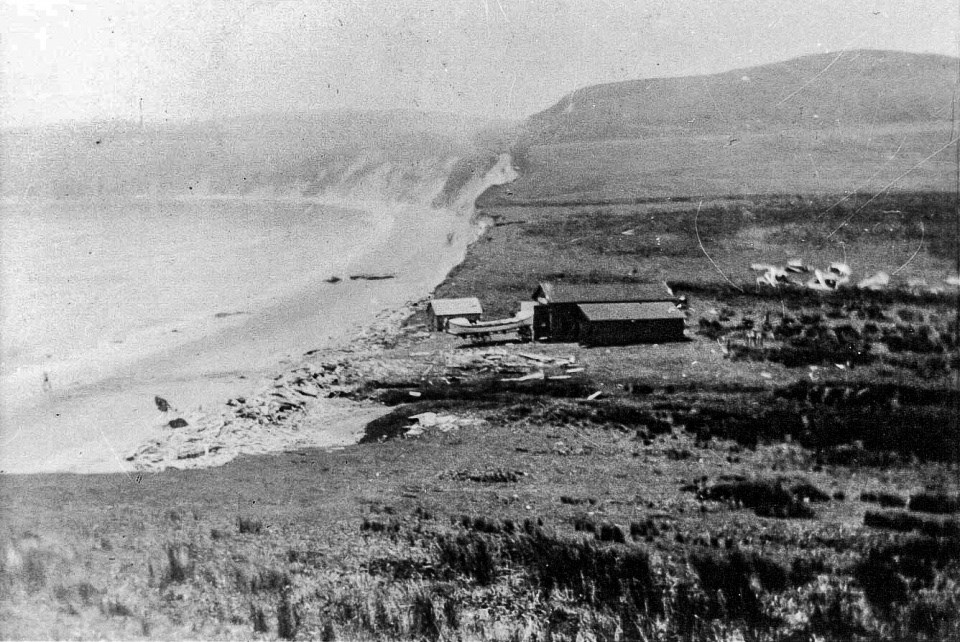 A black and white photo of a garage-like structure and a surf boat adjacent to the beach to the left with cliffs in the background.
