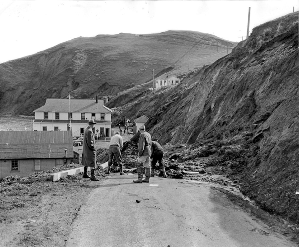 A black and white photo of men clearing landslide debris from a road that leads down to some bay-side buildings. Steep bluffs rise on the right and in the background.