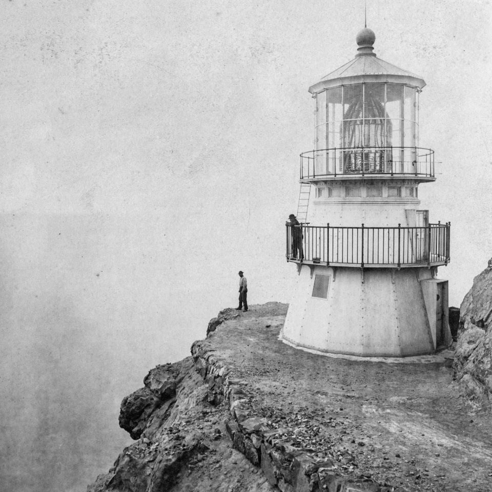 A black and white photo of a short, three-story-tall, white lighthouse on the edge of a cliff. One man stands on the balcony of the tower while a second man stands at the edge of the cliff.