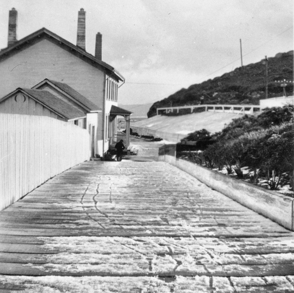A black and white photo of a snowy pathway with tricycle tracks and footprints leading to a two-story house on the left. A man stands near the near corner of the house.