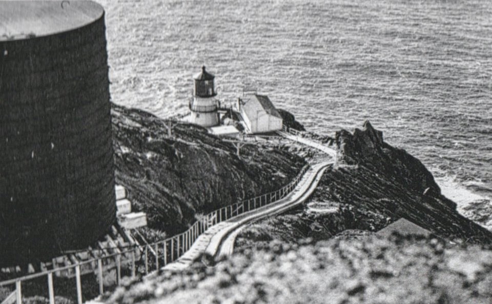 A black and white photo of a three-story-tall lighthouse and two buidlings at the base of wooden stairs. On the left, a large, round, wooden water tank sits at the top of the stairs.