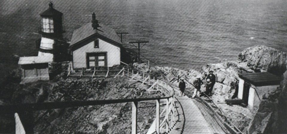 A black and white photo of a wooden ramp running down a rocky, coastal ridge line to a three-story lighthouse tower and two other buildings. A man with three children stand near the base of the stairs.