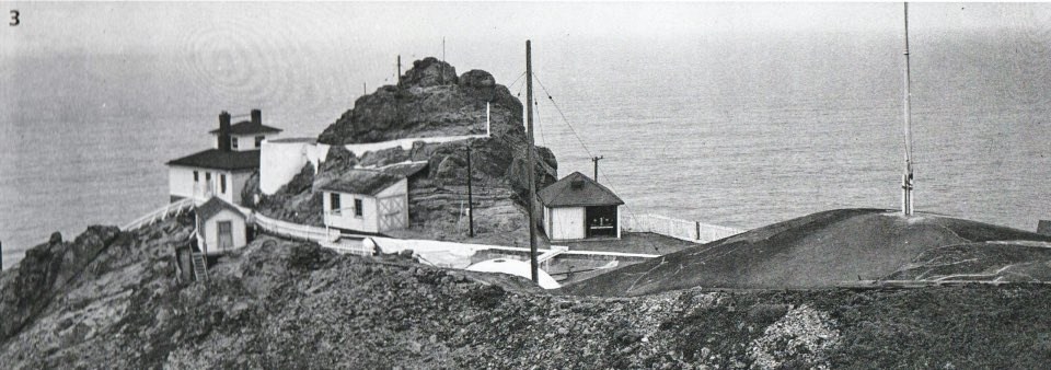 A black and white photo of buildings, rocky outcroppings, and ocean.