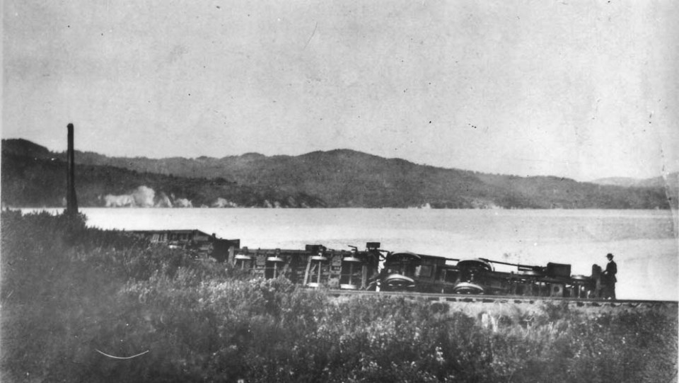 A black and white photo of a train on its side with water and hills in the background.