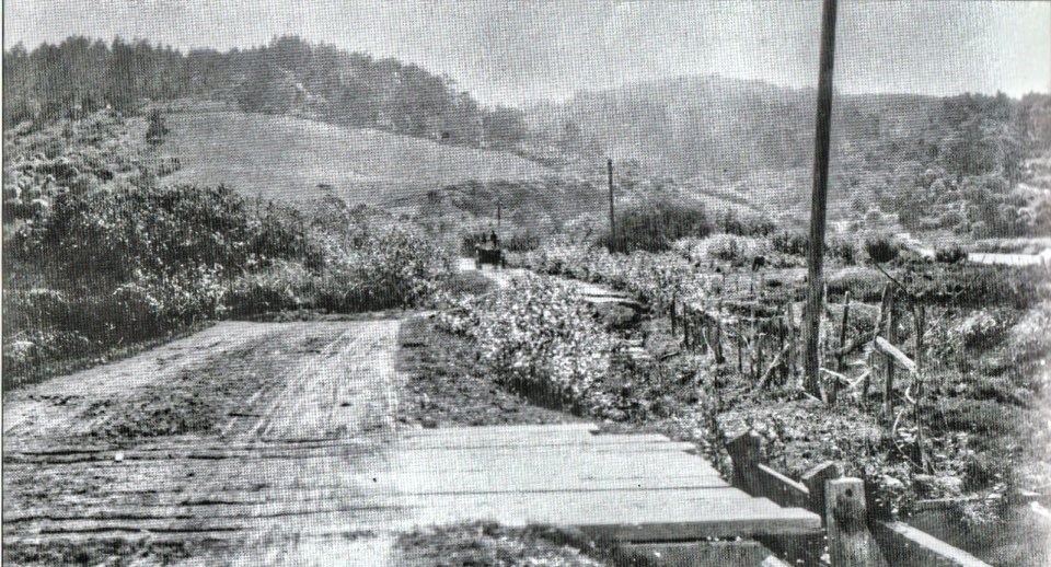 A black and white photo of a road that should be straight but which has an abrupt displacement to the right in the middle distance. An early 1900s automobile is visible beyond where the road is broken..