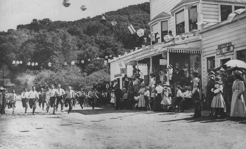 A black and white photo of eleven young men running a race on a dirt road past with a crowd of people and buildings in the early 1900s.