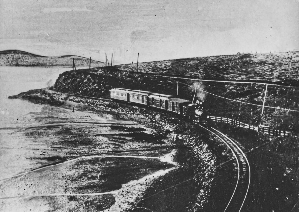 A black and white photo of a steam locomotive pulling four cars on railroad tracks that wind along a bay on the left.