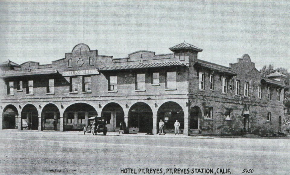 A black and white photo of a two-story brick building with seven ten-foot-tall ground-floor arches along its facade. An early 1900s-era automobile is parked in front of the building. Two men stand in the rightmost archway.